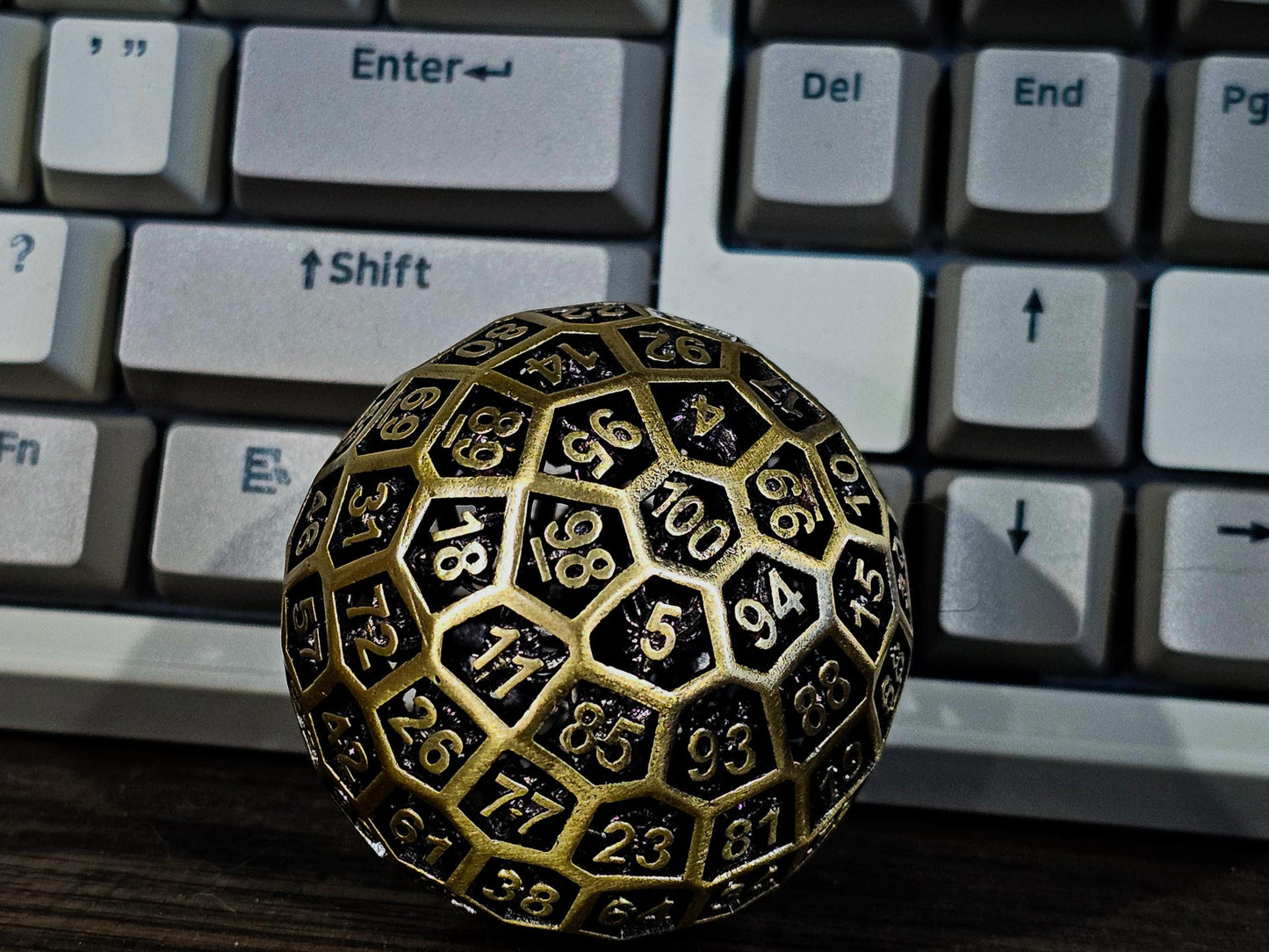 Hollow Metal Ancient Gold D100 DND Dice in front of a white mechanical keyboard that is standing up with its keys facing the camera.