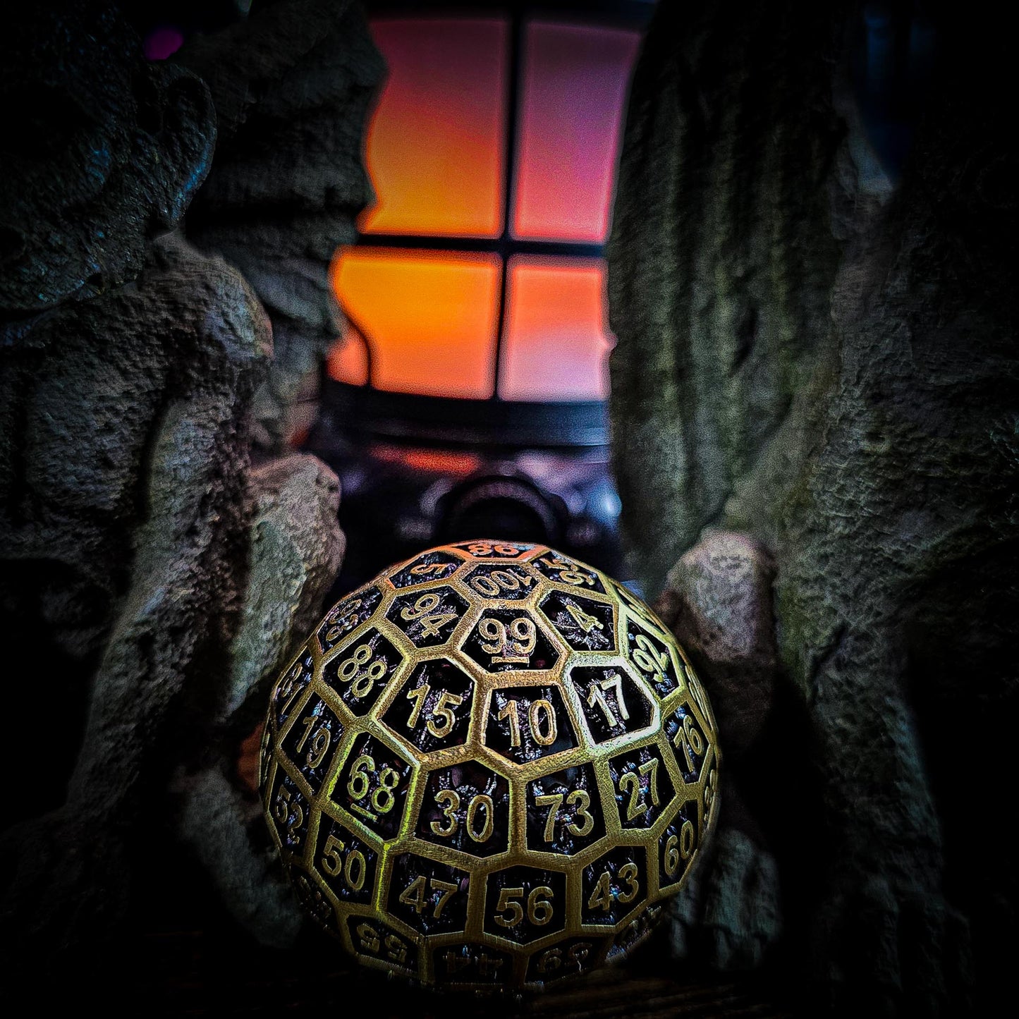 a dark picture of a Hollow Metal Ancient Gold D100 DND Dice between two gargoyles with an orange flickering lamp in the background.