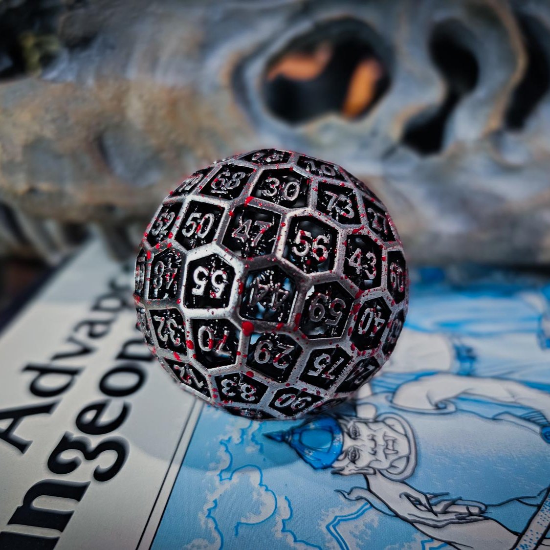silver hollow metal D100 DND Dice with blood spatter sitting on top of a dungeons and dragons manual with a dinosaur skull in the background