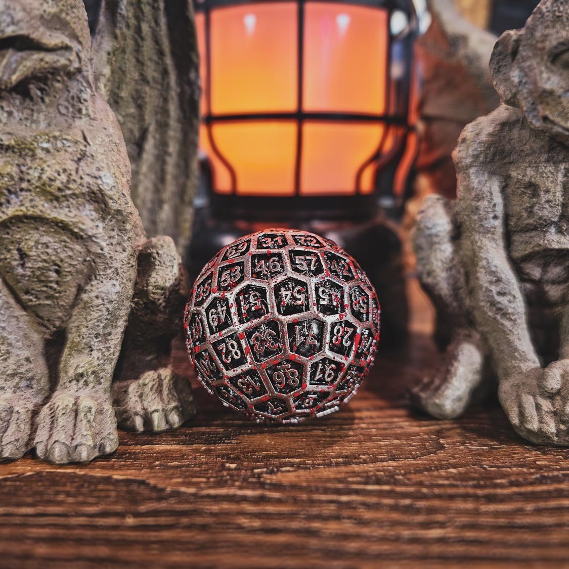 Hollow Metal Silver Blood D100 DND Dice sitting in between two gargoyle statues with an orange flickering lantern in the backdrop.