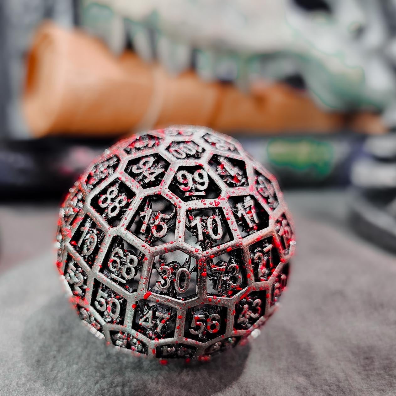 Hollow Metal Silver Blood D100 DND Dice in the foreground with a rolled up tan DND mat and dinosaur skull in the background.