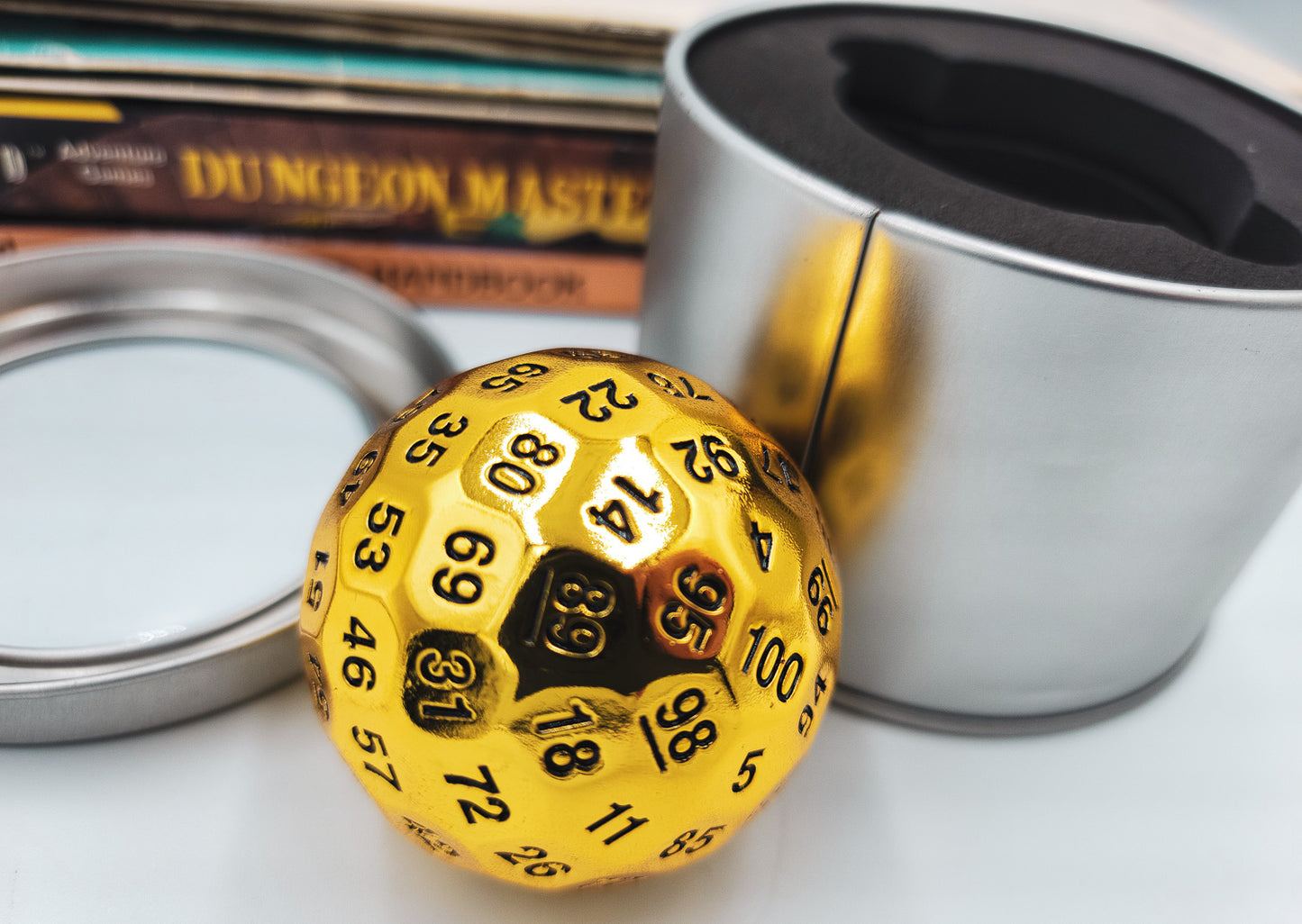 Metal Shiny Gold D100 DND Dice next to a silver metal container and Dungeons and Dragons Books stacked up in the background.