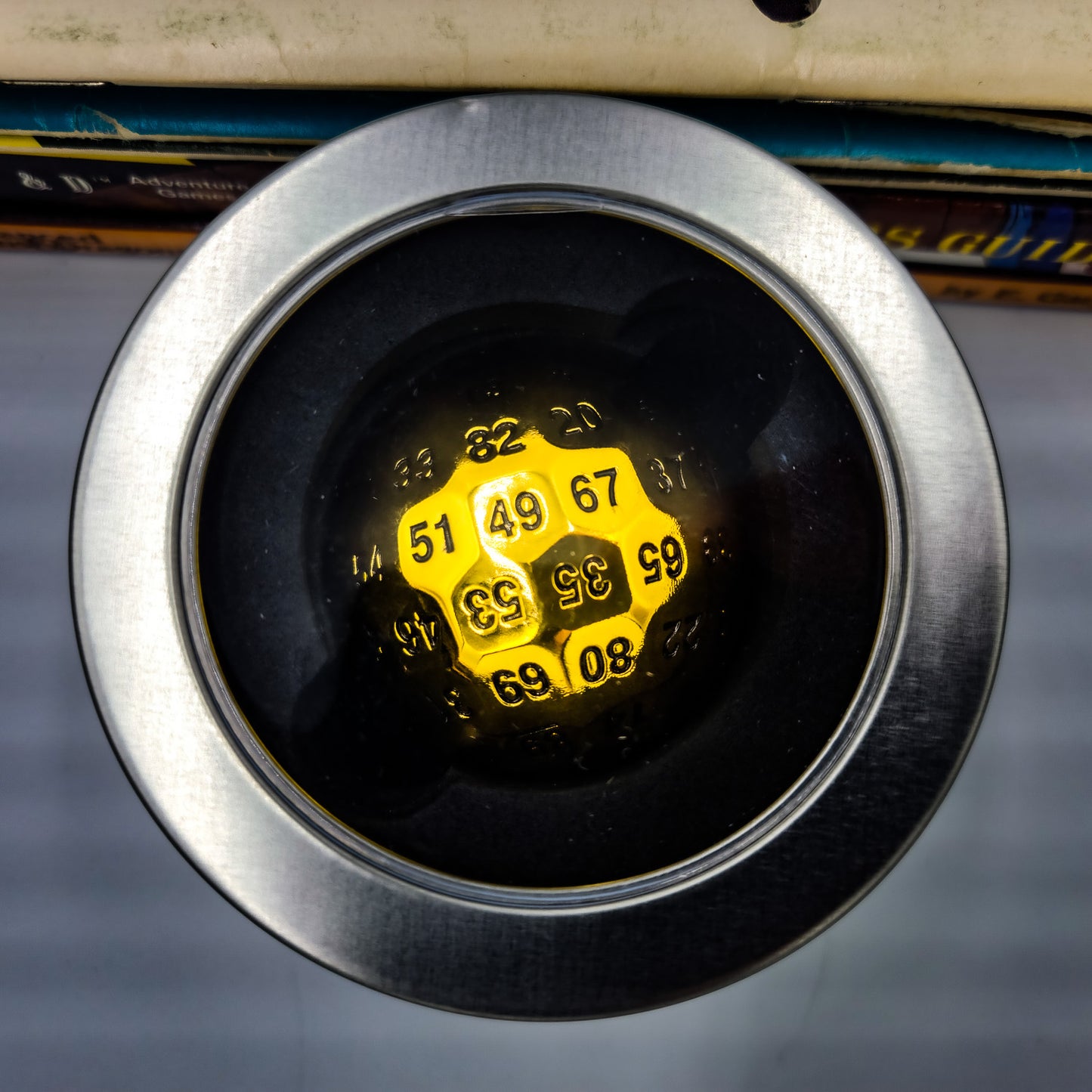 Top down view of a Metal Shiny Gold D100 DND Dice inside a silver metal container with a plastic see through window on the lip.