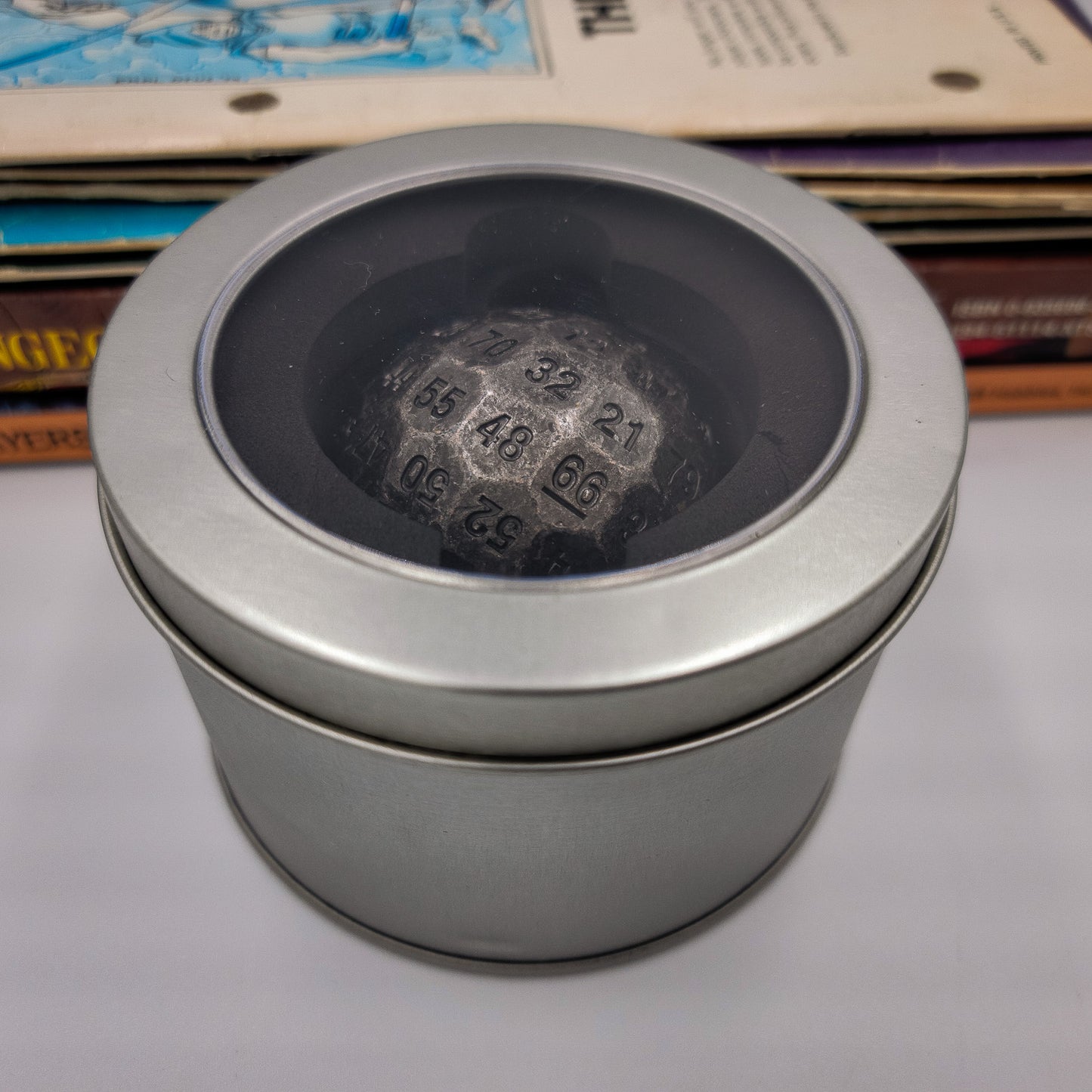 Metal Silver D100 DND Dice inside a silver metal container with plastic see through window on the lid. There is a stack of Dungeons and Dragons manuals behind it.
