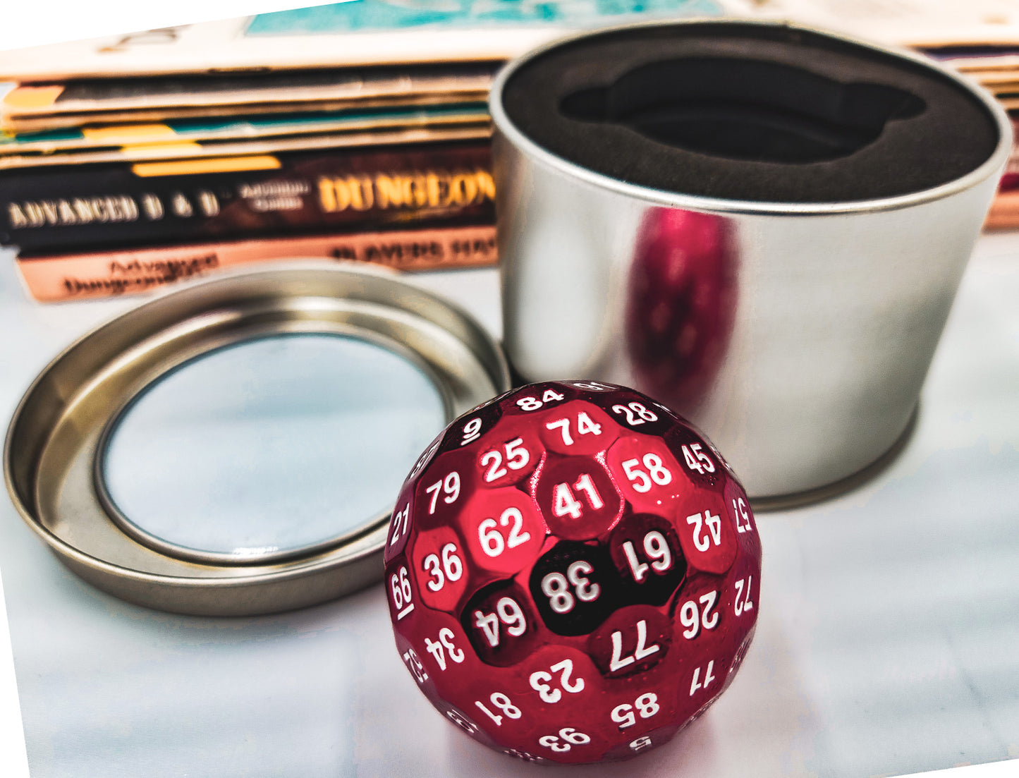 Red Solid Metal D100 DND Dice sitting in front of a silver metal container with its lid removed and a stack of Dungeons and Dragons manuals in the background.