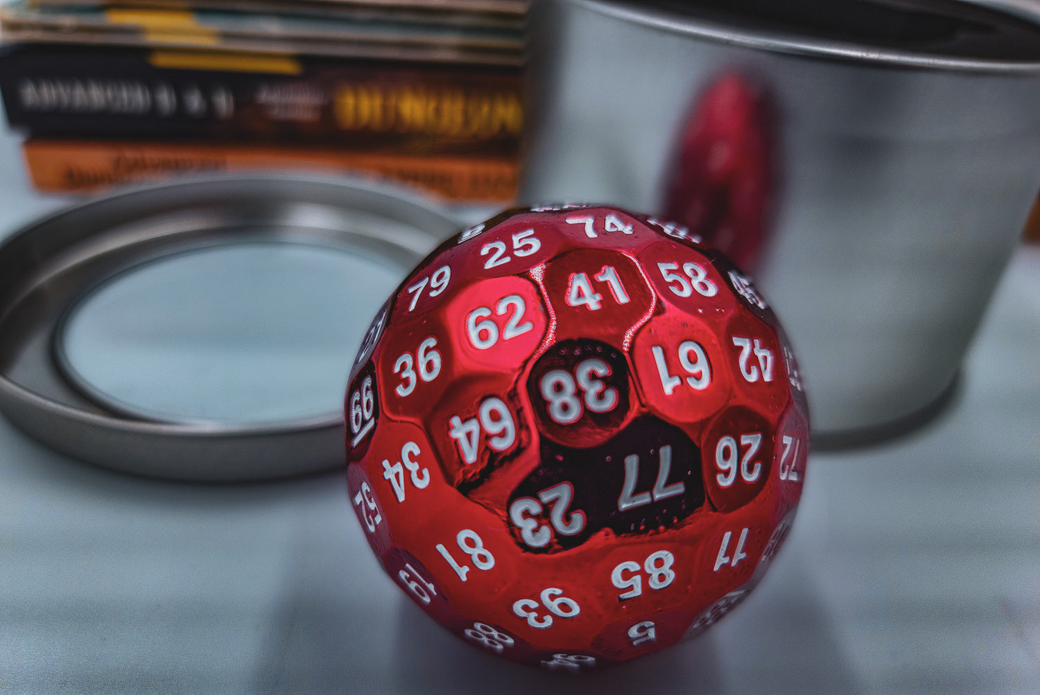 a Red Solid Metal D100 DND Dice sitting in front of a silver metal container and a stack of Dungeons and Dragons booklets.