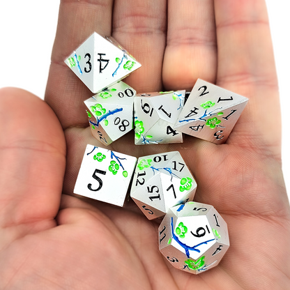 Silver solid metal DND Dice with green japanese plum blossoms and blue twigs etched into the sides laying in the palm of a persons hand.