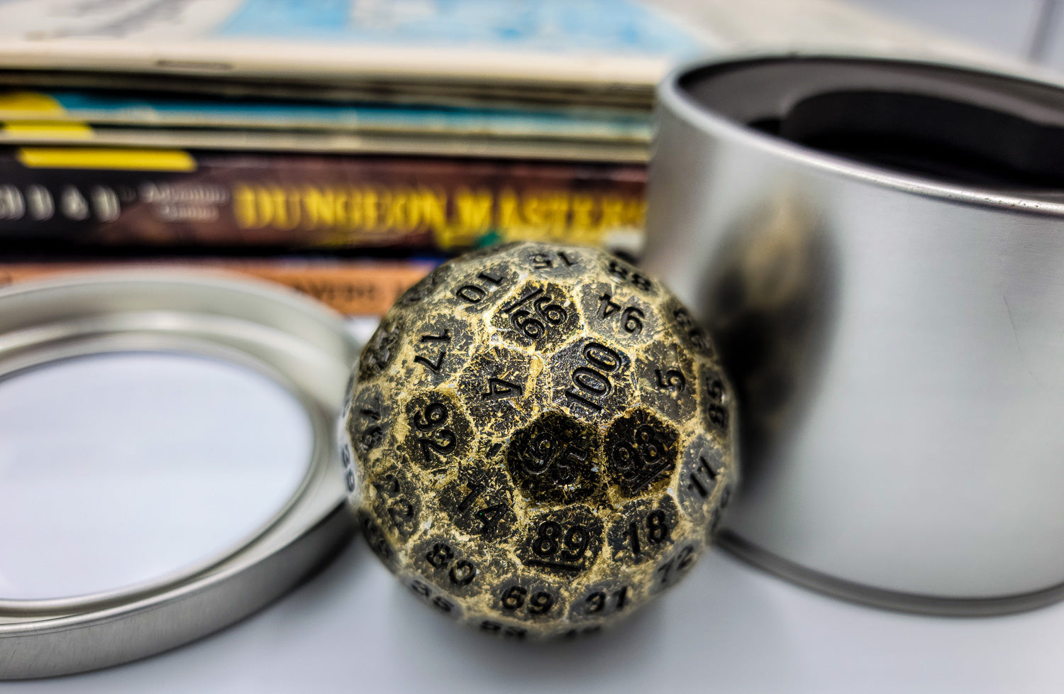 Solid Metal Antique Gold D100 DND Dice in between the silver metal container and lid with a stack of dungeons and dragons booklets behind.