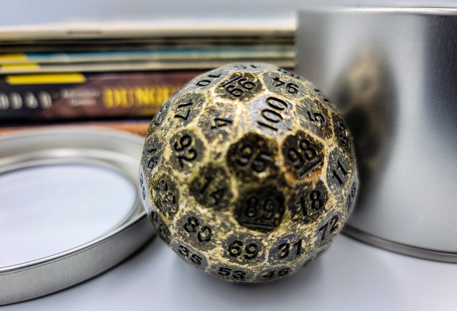 close up shot of Solid Metal Antique Gold D100 DND Dice in between a silver metal container and lid with a stack of dungeons and dragons booklets behind.